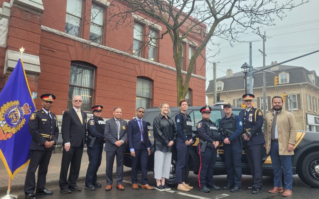 group of eleven people stand in a line in front of a SUV style police vehicle. Brick building in the background and blue flag with police crest to the left of the group.